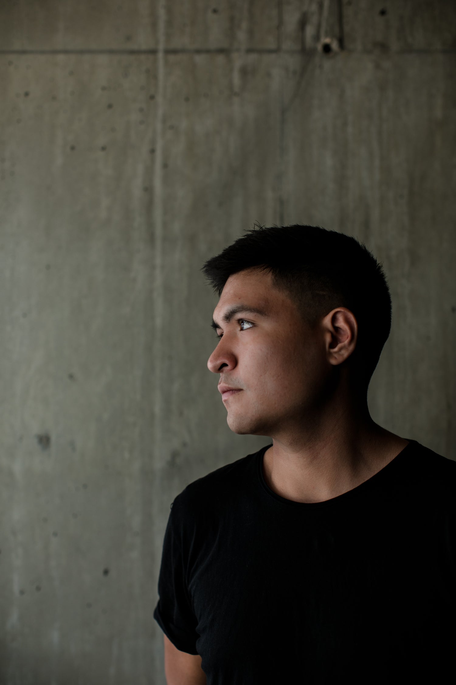 Close-up profile of a young man wearing a black T-shirt, gazing thoughtfully into the distance with a concrete background